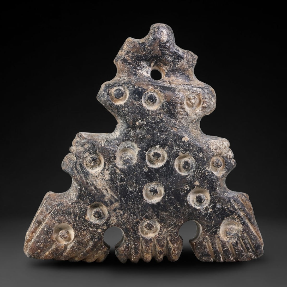 Ancient Elamite Civilization Multi-Figural Circle-and-Dot Stone Amulet (3200-539 BCE), 89AJ: This complex multi-lobed dark stone amulet originates from Elamite Civilization, densely decorated with classic Elamite circle-and-dot motifs interspersed with stylized figural elements. A central upp