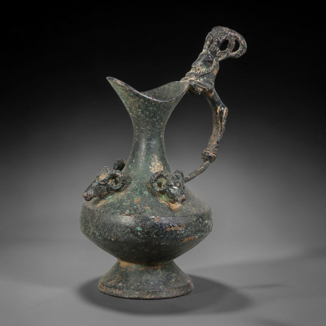 Ancient Seljuk Period Bronze Ewer (c.11th-12th Century CE), 98AK: This ancient Seljuk bronze ewer displays modelled ram-head decorative motifs upon its bulbous body, with an ornate figural-formed handle rising from shoulder to spout. The piece bears deep burial pati