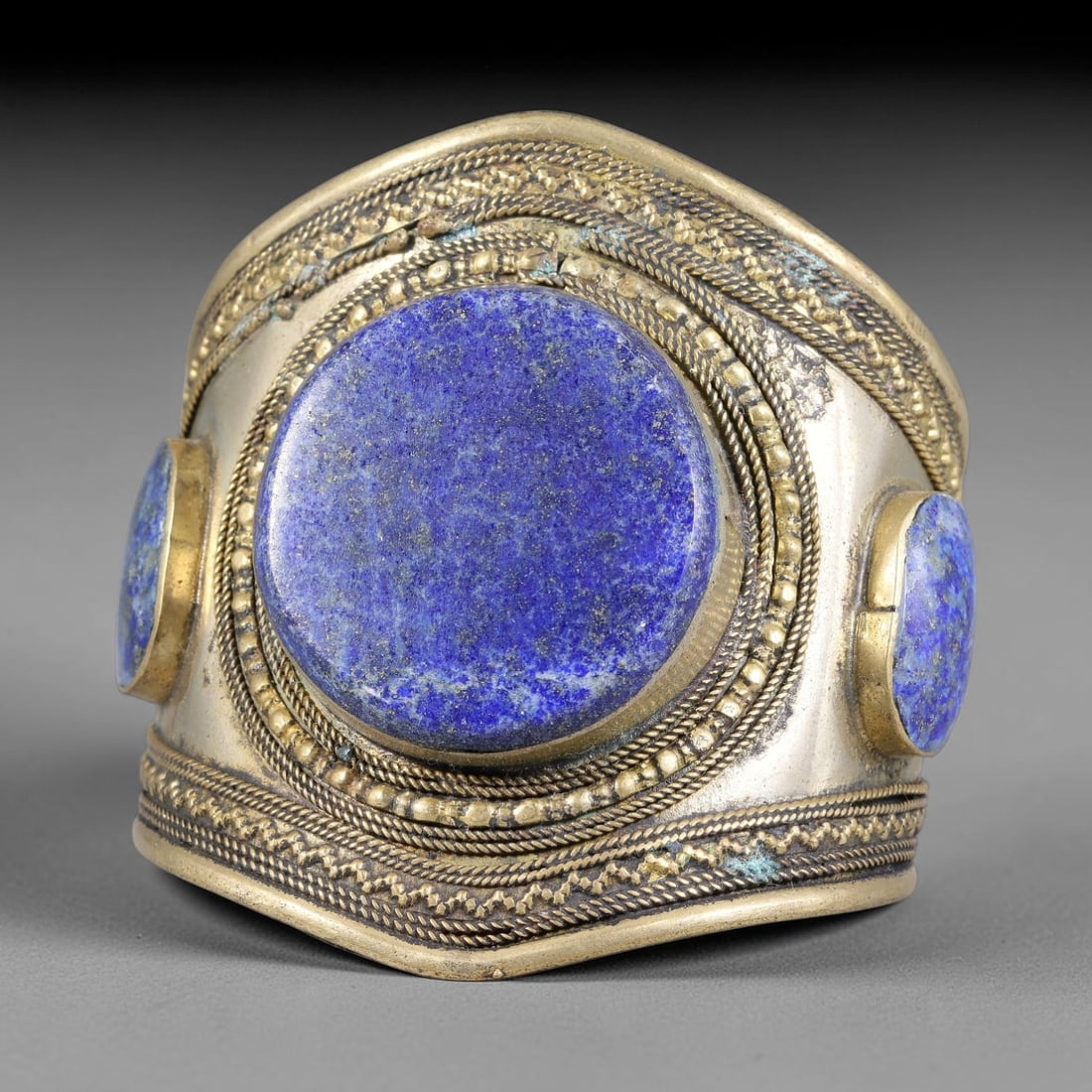 A Central Asian Copper-Alloy Lapis Lazuli Inlaid Cuff Bracelet, 40AK: Wide copper-alloy cuff bracelet set with a large central circular lapis lazuli cabochon flanked by additional stone inlays, all held within heavy bezel mounts. Multiple layers of twisted filigree wire