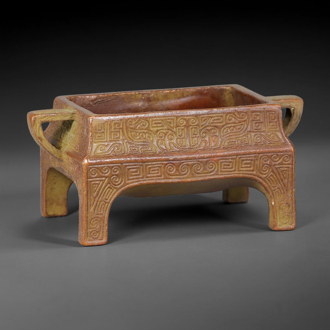 A Chinese Archaic-Style Bronze Rectangular Four-Footed Censer, 89AD: Cast bronze rectangular censer with two looped side handles, standing upon four arched cut-out feet. Exterior walls are densely engraved with interlocking archaic scroll and key-fret decorative patter