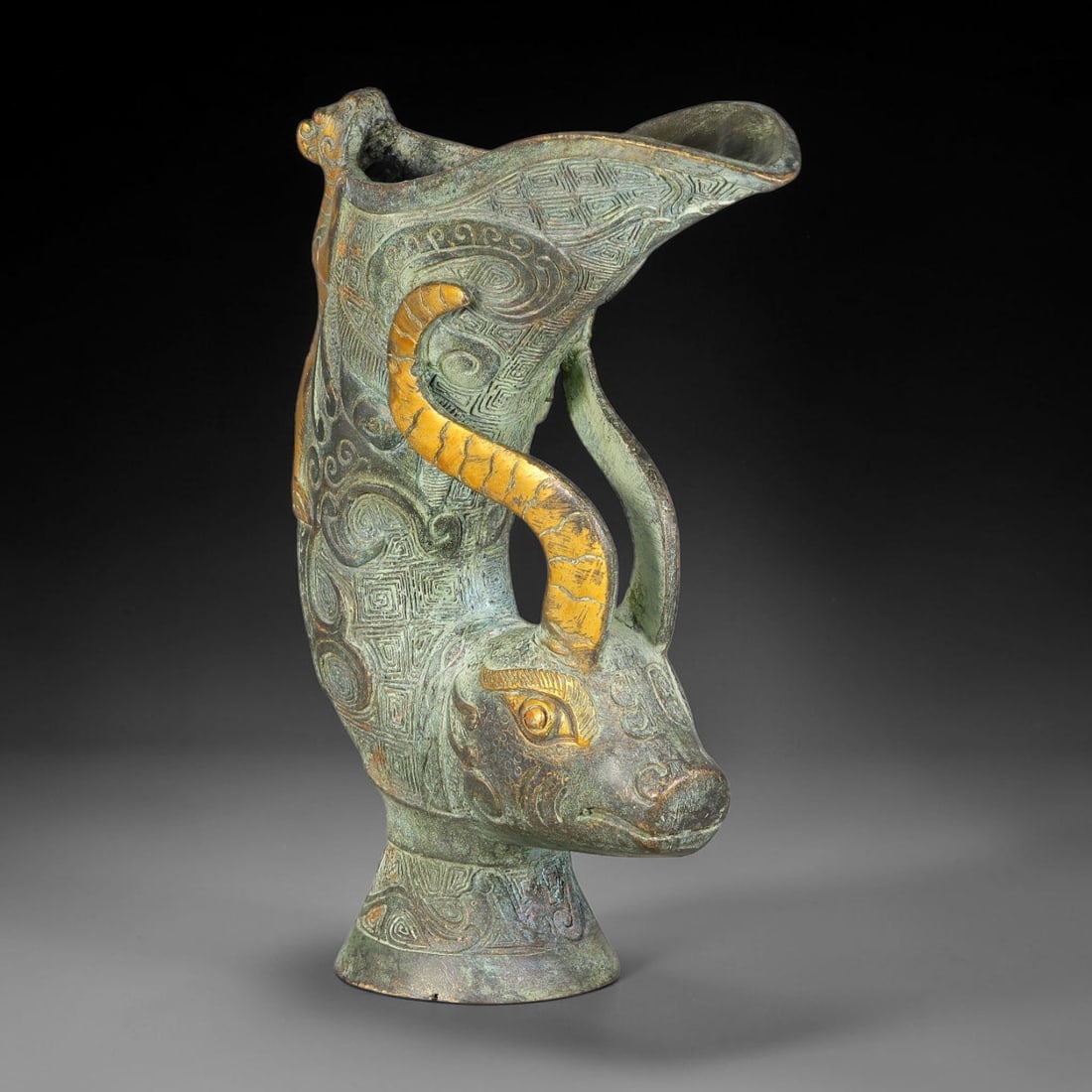A Chinese Archaic-Style Partial Gilt Bronze Ox-Head Rhyton Libation Vessel, 1AJ: Cast bronze rhyton libation vessel modelled with an ox-head forming the lower section, its large gilded curved horn arching upwards to support the flared pouring spout. A small gilded mythical beast c