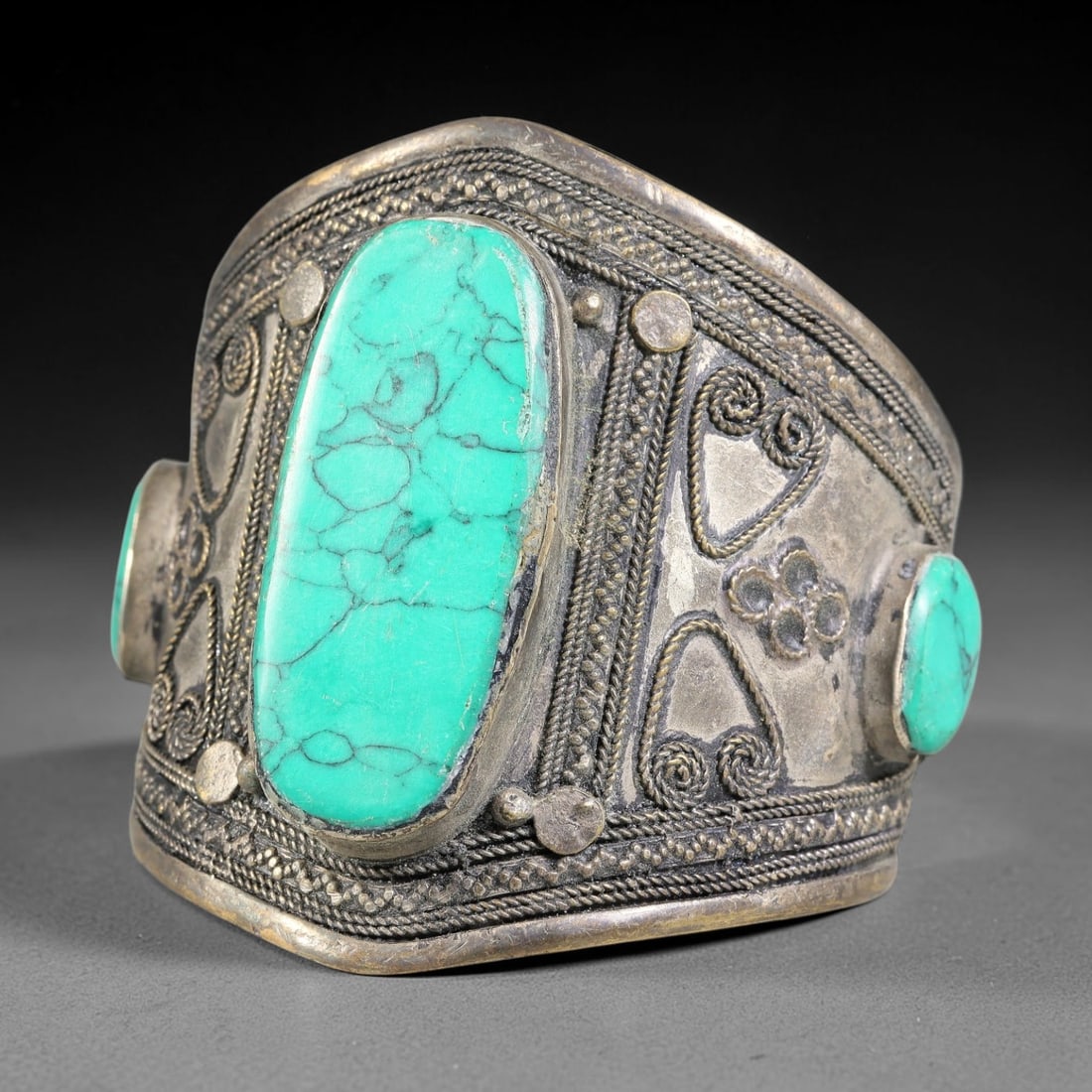 A Central Asian Copper-Alloy Turquoise Inlaid Cuff Bracelet, 36AK: Robust copper-alloy cuff bracelet set with multiple turquoise cabochons within bezel settings. The metal surface features elaborate applied filigree wirework, including scroll and floral-form motifs, 