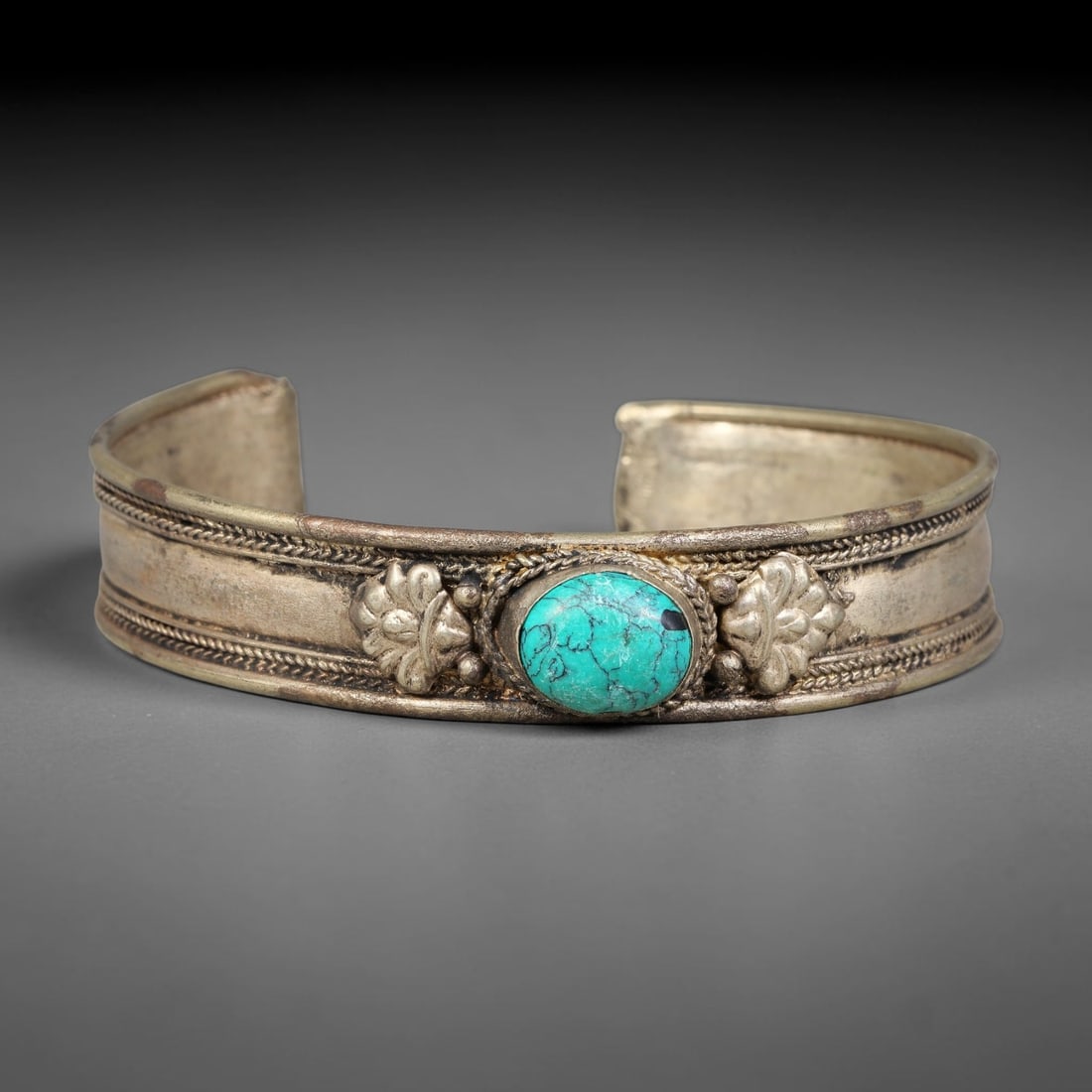 A Central Asian Copper-Alloy Turquoise Inlaid Open-Cuff Bracelet, 49AK: Open-form copper-alloy cuff bracelet centering an oval turquoise cabochon held within a twisted-wire bezel mount. Repousse floral relief ornaments sit on either side of the stone, paired with small sp