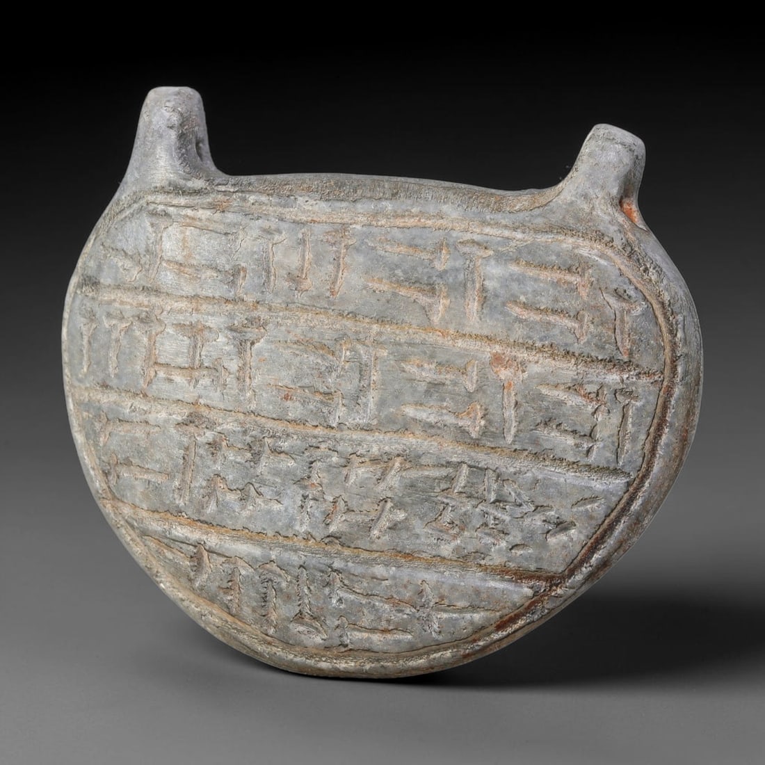 Ancient Uruk Period Hard-Stone Circular Inscribed Apotropaic Amulet (c.3500-3100 BCE), 63AJ: Circular-form apotropaic amulet of the Uruk cultural tradition, fitted with dual integrated suspension lugs at the upper margin for personal wear. Its face bears multiple horizontal registers of incis