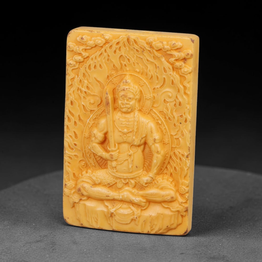 A Chinese Acala Deity Carved Tagua Nut Pendant Amulet, 90BC: Carved from tagua nut, this rectangular tablet-form amulet is rendered in high relief. Acala, the immovable protective guardian, is depicted seated upon a rocky base within full lotus posture, graspin