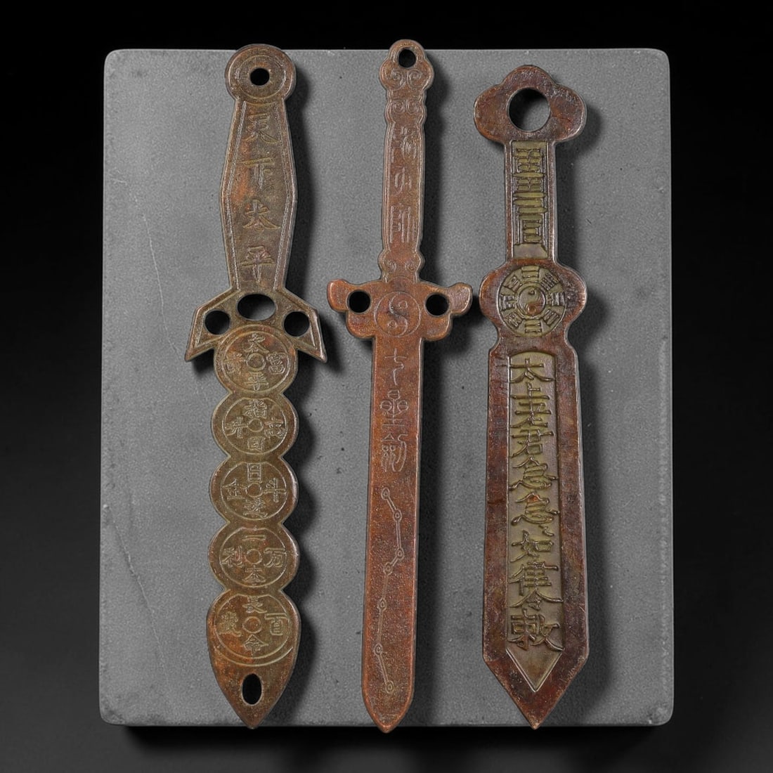 Lot of Three Chinese Bronze Taoist Ritual Talisman Sword Amulets, 34AD: Cast in bronze, this group of three flat-bodied ritual sword amulets are pierced with suspension holes for hanging. Each piece bears deeply incised Taoist iconography: bagua trigram emblem, yin-yang s