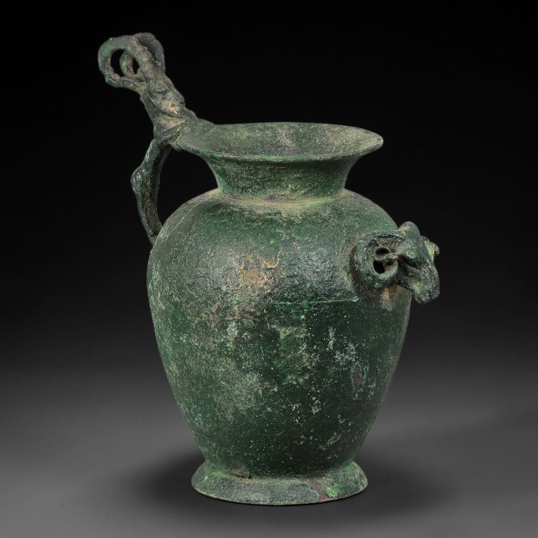 Ancient Seljuk Period Bronze Zoomorphic Vessel (c.1037-1194 CE), 36AL: Ancient Seljuk period bronze vessel, featuring a sculpted ram head spout and ornate zoomorphic handle, retaining deep stable green burial patina across the body. Dimensions measure 13.5 x 9 cm.