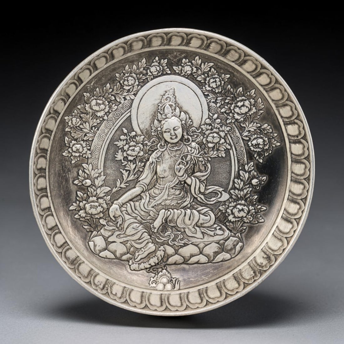 A Tibetan Tara Figure German Silver Decorative Plate, 31AD: A circular plate in German silver (a nickel-silver alloy), the center decorated in high relief with a Tara figure. The deity is shown seated in a meditative posture upon a lotus throne, surrounded by 