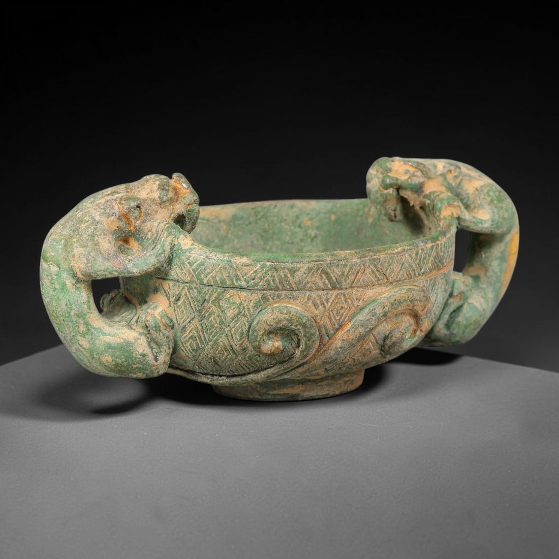 A Chinese Bronze Twin Dragon-Handled Ritual Bowl, 60AD: Cast in bronze, this shallow ritual bowl features two sculpted dragon-form handles gripping the vessel rim from either side. The exterior surface is decorated with finely incised geometric and scroll 