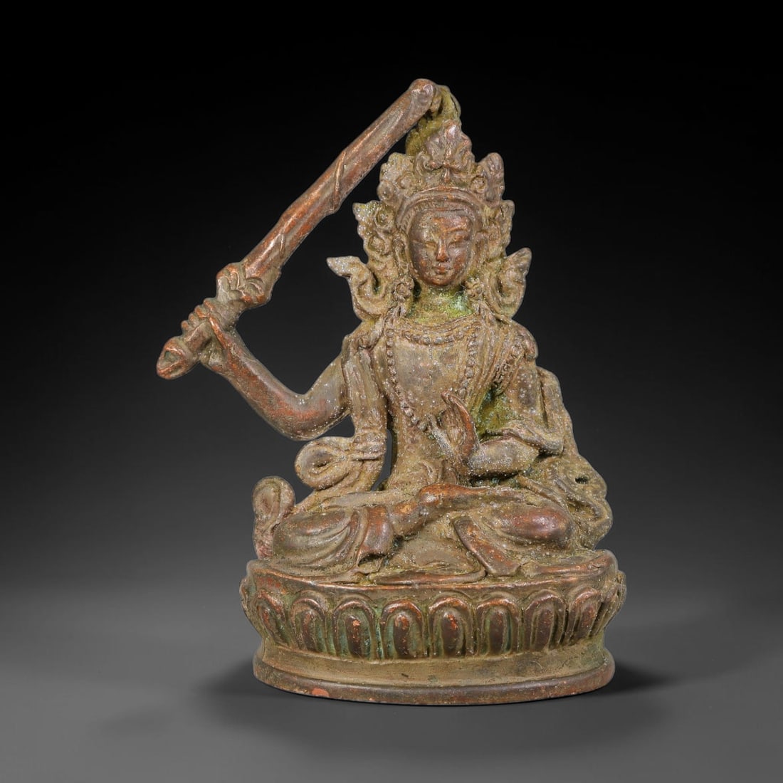 A Tibetan Bronze Manjushri Seated Sculpture Figurine, 40AD: Cast in bronze, this effigy depicts Manjushri seated in meditative lotus position upon a detailed double-petal lotus base. The deity holds the flaming wisdom sword aloft in one hand, while the other f