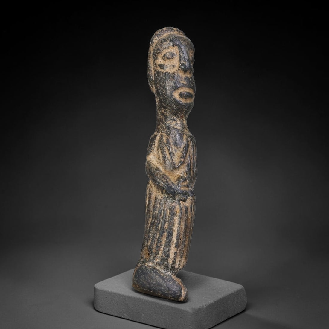 Ancient Elamite Civilization Standing Anthropomorphic Stone Votive Figurine (3200-539 BCE), 8AK: This tall carved dark stone votive figurine originates from Elamite Civilization, depicting a standing human figure with stylized facial features and incised linear detailing rendering draped garments