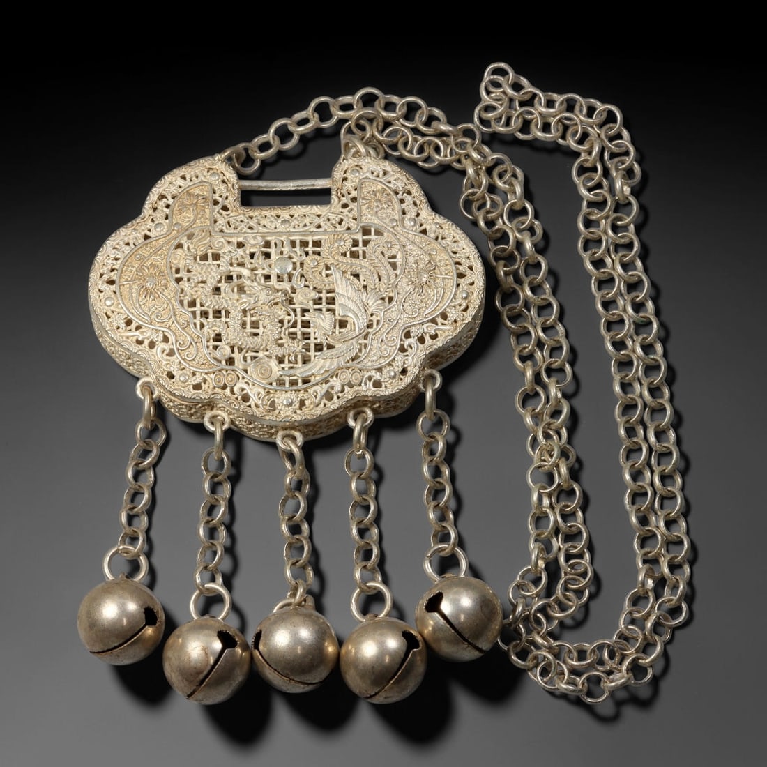 A Chinese German Silver Openwork Ruyi Lock Pendant with Bells, 64AV: Ruyi-shaped German silver lock pendant crafted with intricate double-sided openwork relief. One side depicts figural scene within architectural interior, the reverse features dragon and phoenix motif 