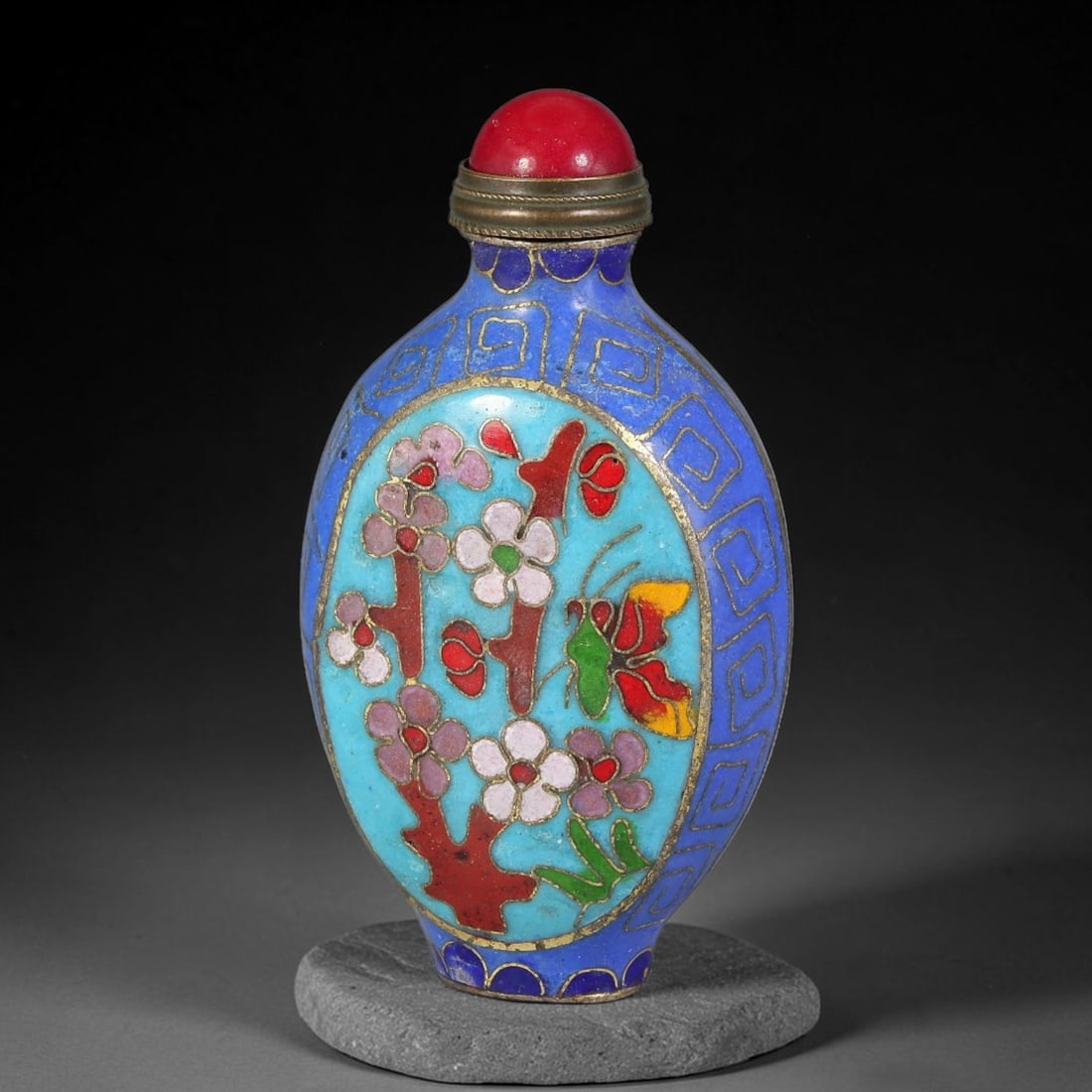 A Chinese Cloisonne Enamel Snuff Bottle with Plum Blossom and Butterfly Motif, 1AV: Flattened oval snuff bottle executed in cloisonne enamel. The deep blue ground is decorated with geometric key-fret border patterns framing two large pale turquoise enamel medallions. Each medallion f