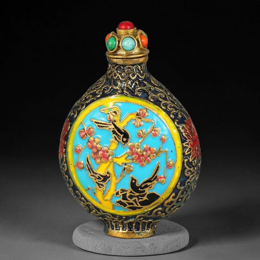 A Chinese Gilt Metal Enamel Snuff Bottle with Magpie and Plum Blossom Motif, 33AV: Flat circular snuff bottle crafted in gilt metal, fitted with a stopper set with simulated coral, turquoise and orange cabochons. The exterior features intricate gilt scrolling patterns on a dark grou