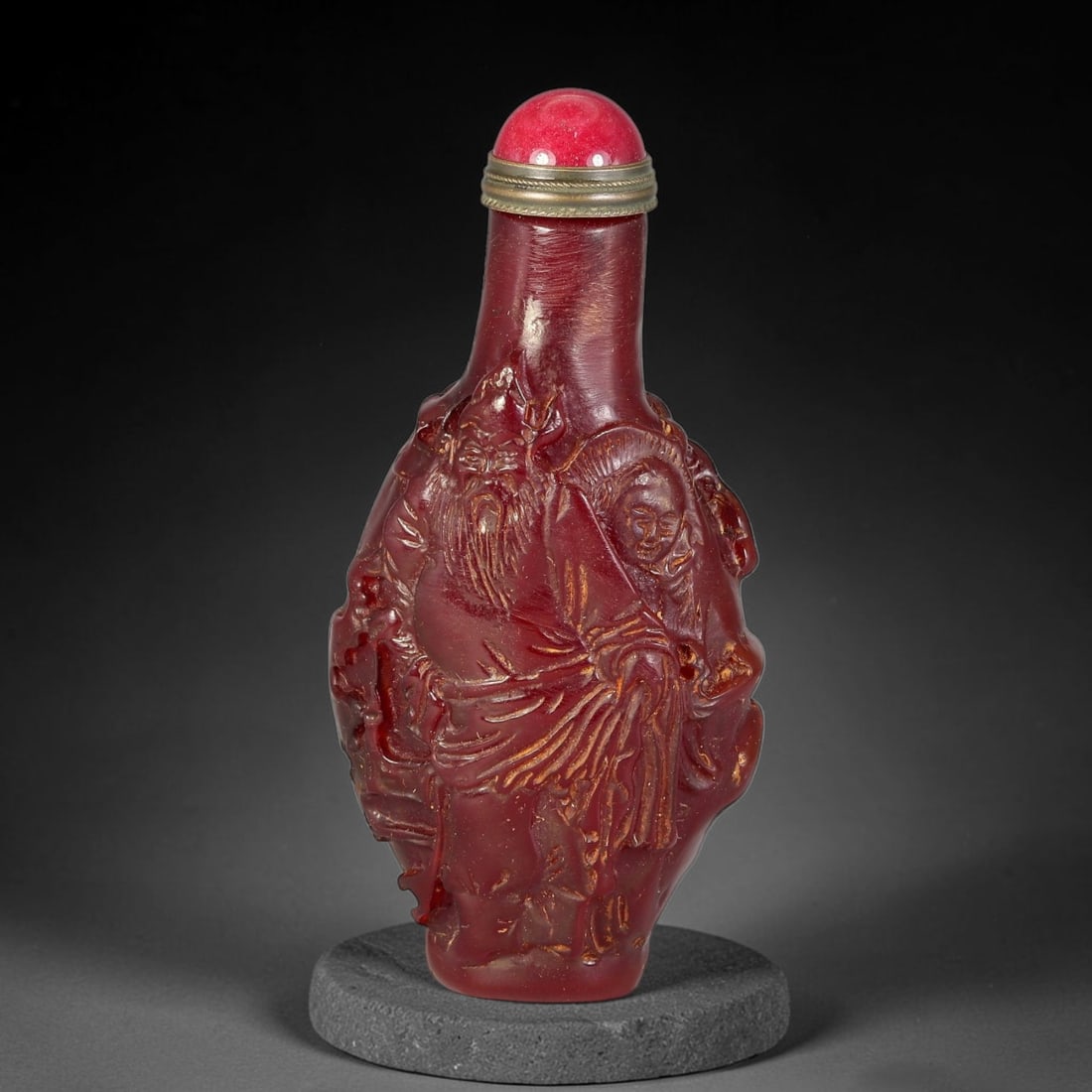 A Chinese Red Amber Carved Figural Snuff Bottle, 10AV: Deep red amber snuff bottle with high-relief carved decoration on both sides. One face depicts a bearded elder and attendant in traditional robes, framed by naturalistic rockwork; the reverse features