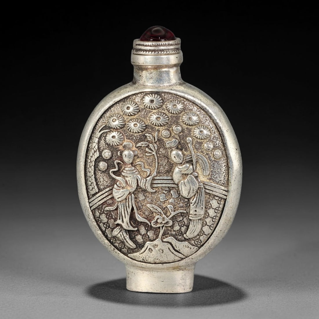 A Chinese Repousse Metal Ladies Figural Snuff Bottle, 63AV: Moon flask shaped snuff bottle decorated in raised repousse relief depicting two robed female figures within a garden setting beneath flowering chrysanthemums, set upon a textured stippled background.