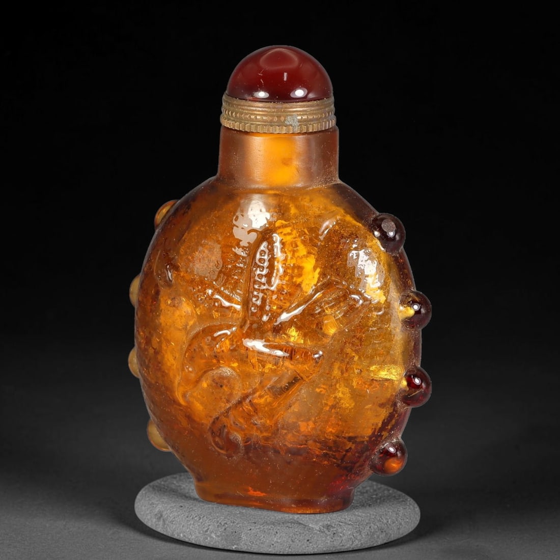 A Chinese Amber Glass Relief Snuff Bottle, 50AV: Flattened ovoid snuff bottle crafted from translucent amber glass with internal speckled inclusions. The exterior features raised relief carving and rows of protruding bead bosses along the outer peri