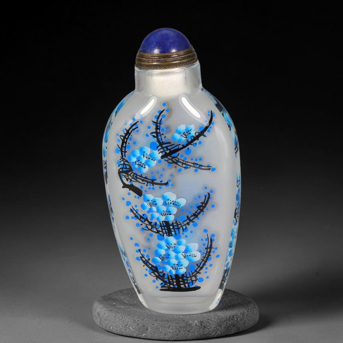 A Chinese Inside Painted Glass Plum Blossom Snuff Bottle, 60AV: Slender clear glass snuff bottle finely decorated with interior painting of plum blossom branches rendered in vibrant blue ink tones against a pale frosted ground. It is fitted with a deep blue domed 