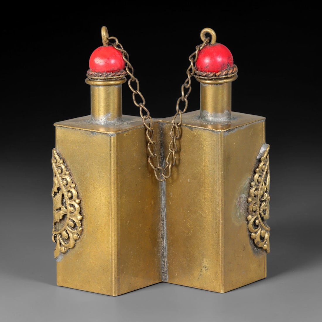 A Chinese Conjoined Twin Brass Snuff Bottle, 67AV: Conjoined twin rectangular brass snuff bottle, formed by two adjoining square vessels. Each neck is fitted with a red spherical stopper secured by a small brass connecting chain. Ornate raised foliate