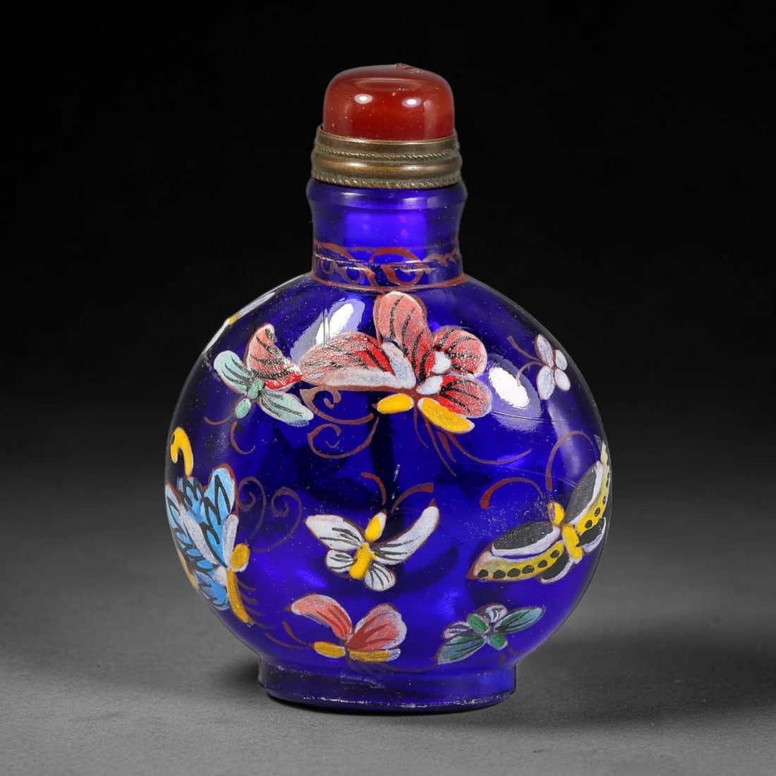 A Chinese Cobalt Blue Glass Enamel Butterfly Snuff Bottle, 46AV: Globular translucent cobalt blue glass snuff bottle decorated with lively polychrome enamel butterflies and delicate scrolling vine motifs across the surface. The neck is adorned with fine reddish-bro