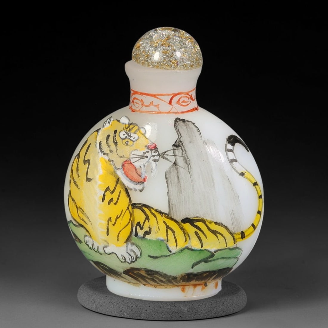 A Chinese White Glass Enamel Painted Tiger Snuff Bottle, 3AV: Globular opaque white glass snuff bottle decorated with hand enamel painting of a roaring tiger resting amid rocks and foliage. The neck features delicate red scroll ornamentation, topped by a gold-fl