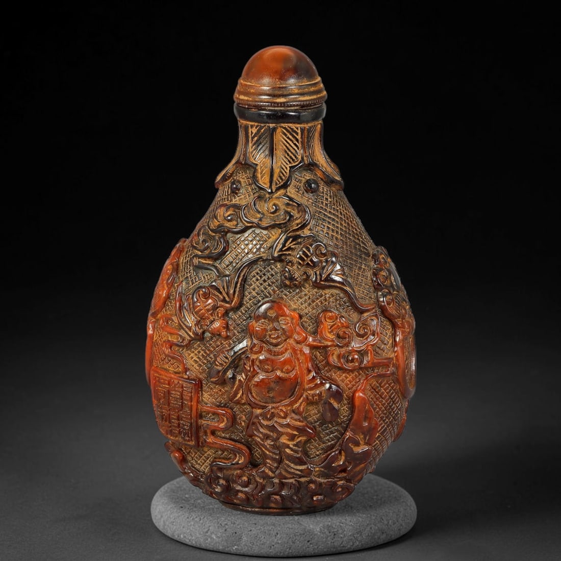 A Chinese Amber Resin Deeply Carved Snuff Bottle, 70AV: Pear-shaped snuff bottle crafted from amber resin, finely carved in deep relief featuring Budai, bat motifs, cloud scrolls and geometric diaperwork across the body. The shoulder displays stylized leaf