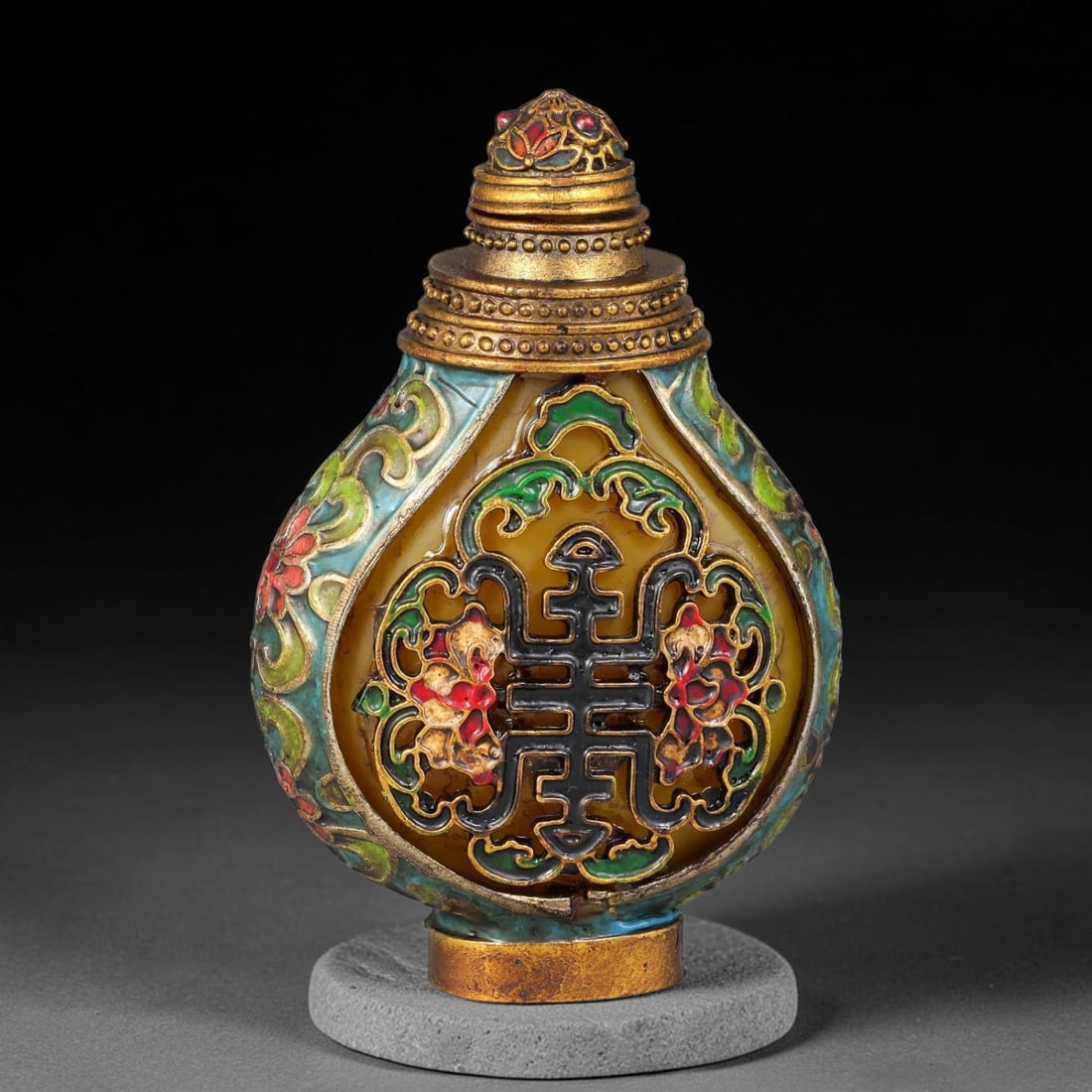 A Chinese Gilt Metal Enamel Overlay Snuff Bottle with Longevity Character Openwork, 34AV: Pear-shaped snuff bottle with yellow translucent panel base, topped with a multi-tiered gilt stopper decorated with enamel details. The front features elaborate polychrome enamel openwork centred on t