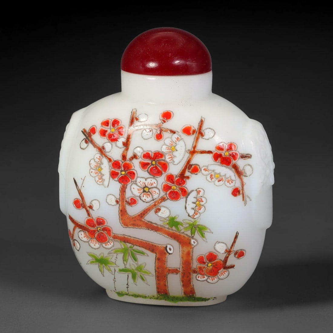 A Chinese White Peking Glass Enamel Plum and Bamboo Snuff Bottle, 98AV: Ovoid white Peking glass snuff bottle with moulded mask handles at the shoulders, decorated with polychrome enamel flowering plum branches paired with bamboo. It is topped by a smooth solid red domed 