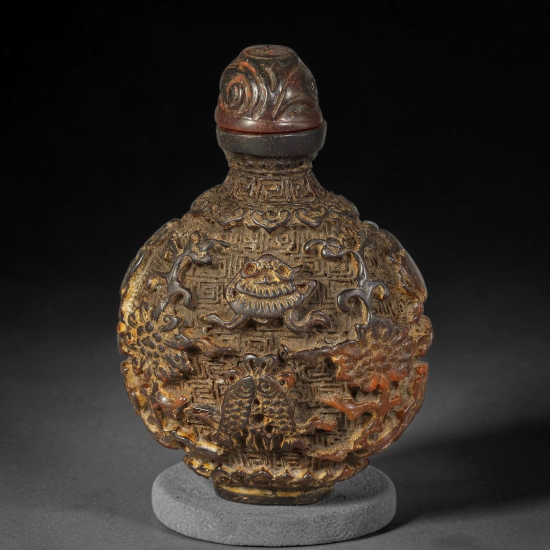 A Chinese Carved Amber Resin Snuff Bottle with Auspicious Symbols, 91AL: Flattened globular snuff bottle densely carved in high relief with traditional auspicious motifs including mythical creature and paired fish, set against a geometric patterned background accented with