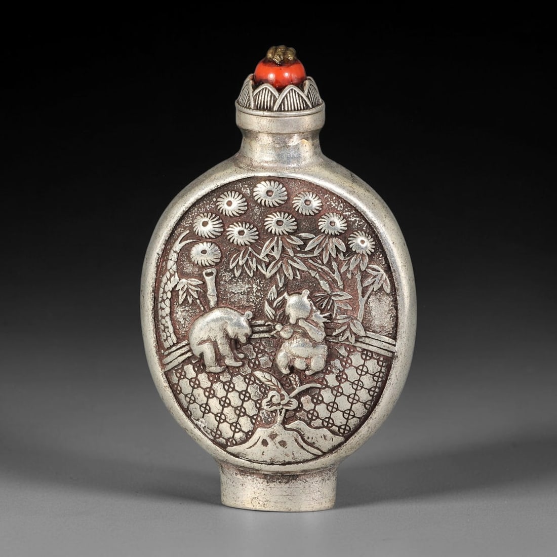 A Chinese Repousse Metal Panda Snuff Bottle, 62AV: Moon flask shaped snuff bottle with raised repousse decoration depicting two pandas amid bamboo and chrysanthemum blossoms over a geometric lattice ground. It is fitted with a red bead stopper, and be