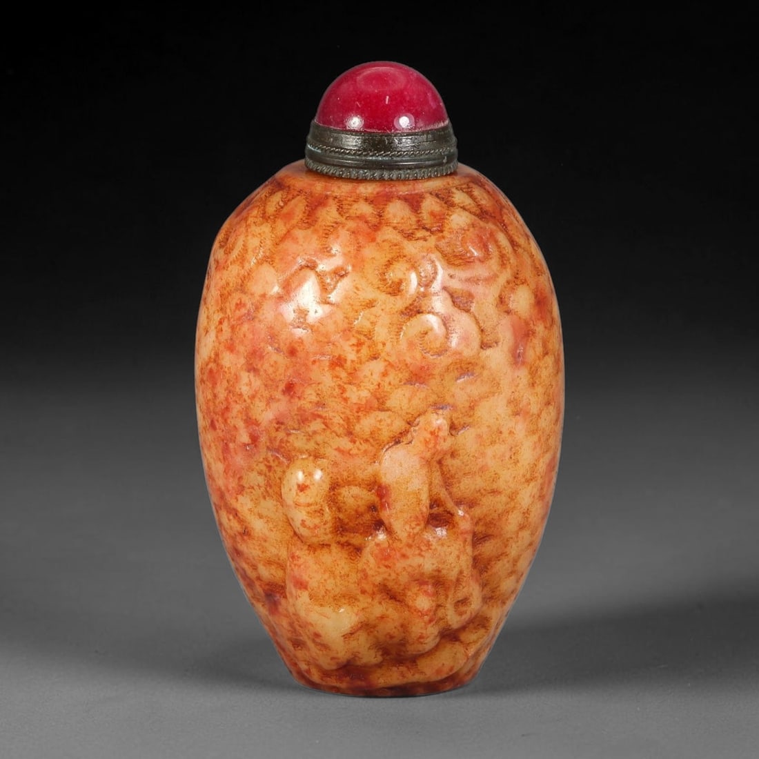 A Chinese Jade Relief Carved Snuff Bottle, 56AV: Ovoid snuff bottle carved from naturally mottled warm-toned jade, decorated with raised relief figural and scroll ornamentation across the body. It is fitted with a deep red domed stopper secured by a