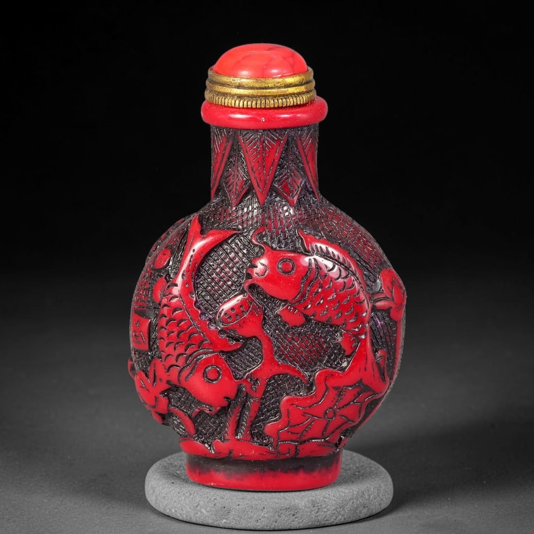 A Chinese Red Carved Koi and Lotus Snuff Bottle, 5AX: Globular snuff bottle finely carved with koi fish swimming among lotus foliage over a textured patterned background, the neck decorated with stylized leaf motifs. It is mounted with a red domed stoppe