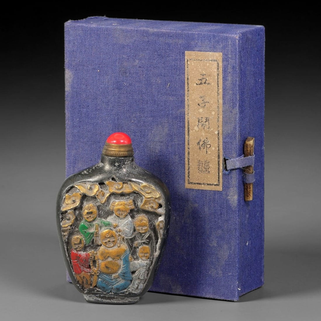 A Chinese Painted Relief Carved Laughing Buddha Snuff Bottle with Original Box, 74AV: Pear-shaped snuff bottle of dark toned material, decorated with polychrome raised relief carving depicting a laughing Buddha surrounded by boys, with cloud scrolls and ornamental borders at the upper 