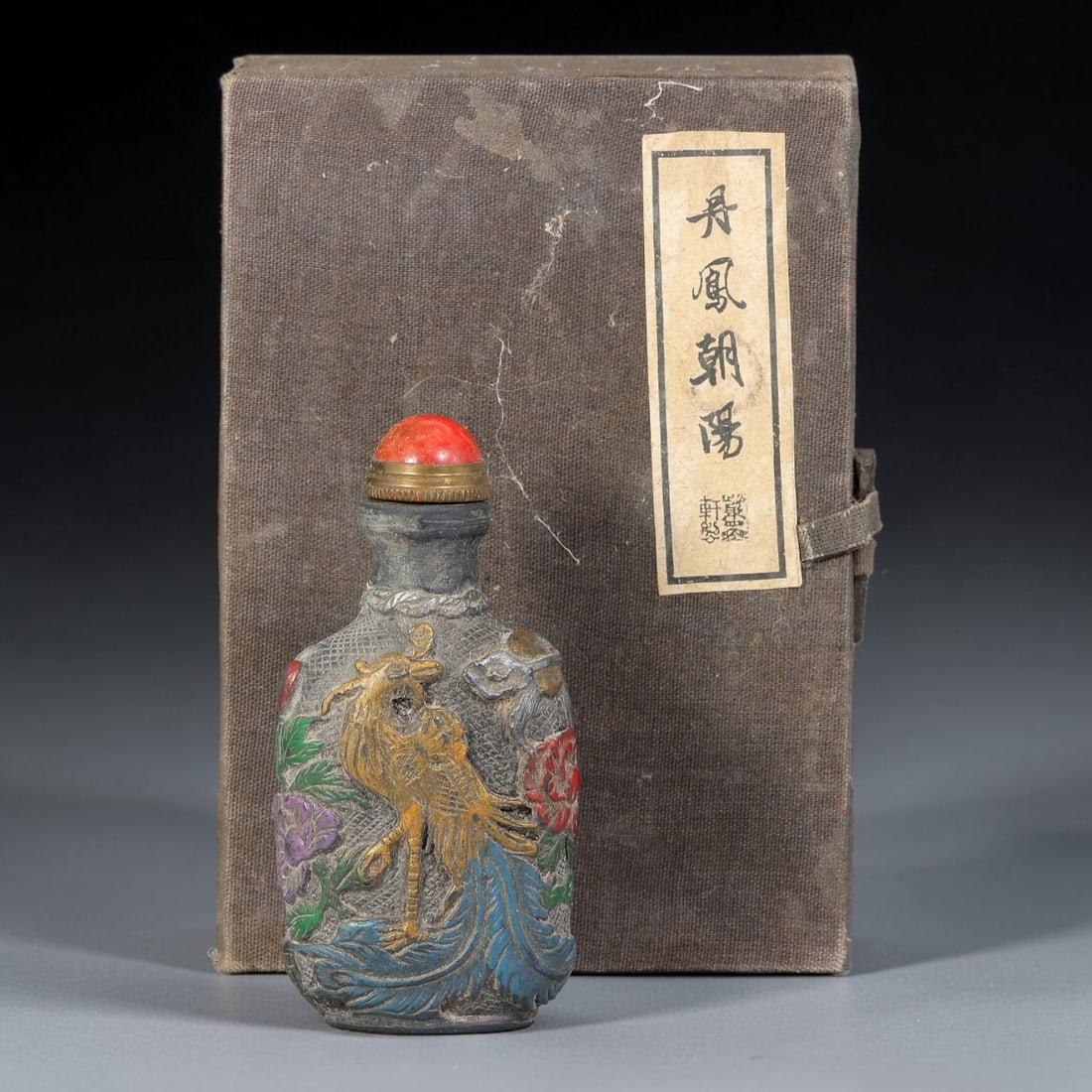 A Chinese Painted Relief Carved Phoenix Snuff Bottle with Original Box, 77AV: Rectangular snuff bottle of dark toned material, decorated with polychrome raised relief carving, one side depicting a phoenix amid peonies and foliage, the reverse with two phoenixes in flight beside