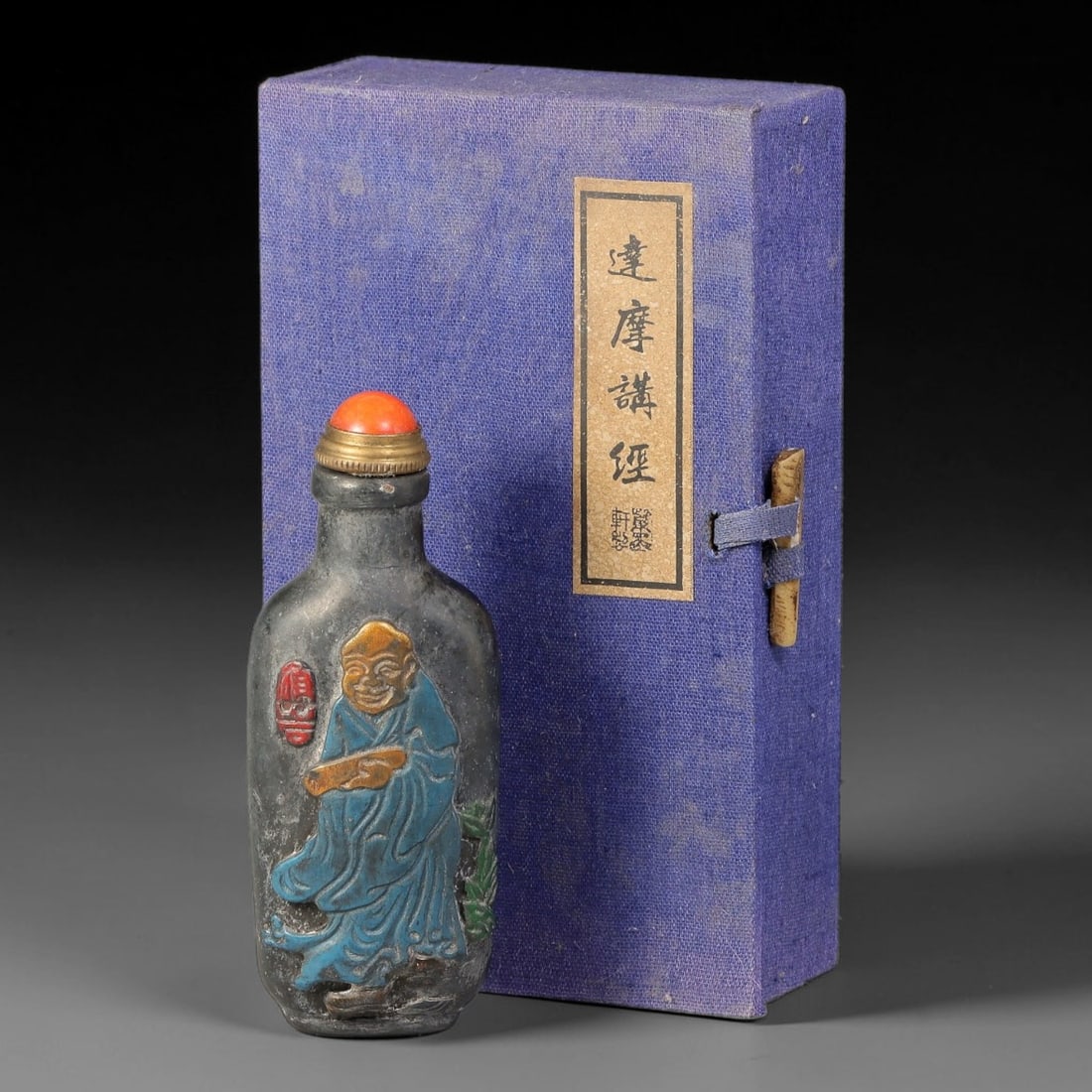 A Chinese Painted Relief Carved Bodhidharma Snuff Bottle with Original Box, 75AV: Rectangular snuff bottle of dark toned material, decorated with polychrome raised relief carving, one side depicting Bodhidharma preaching sutras in coloured robes, the reverse showing Bodhidharma cro