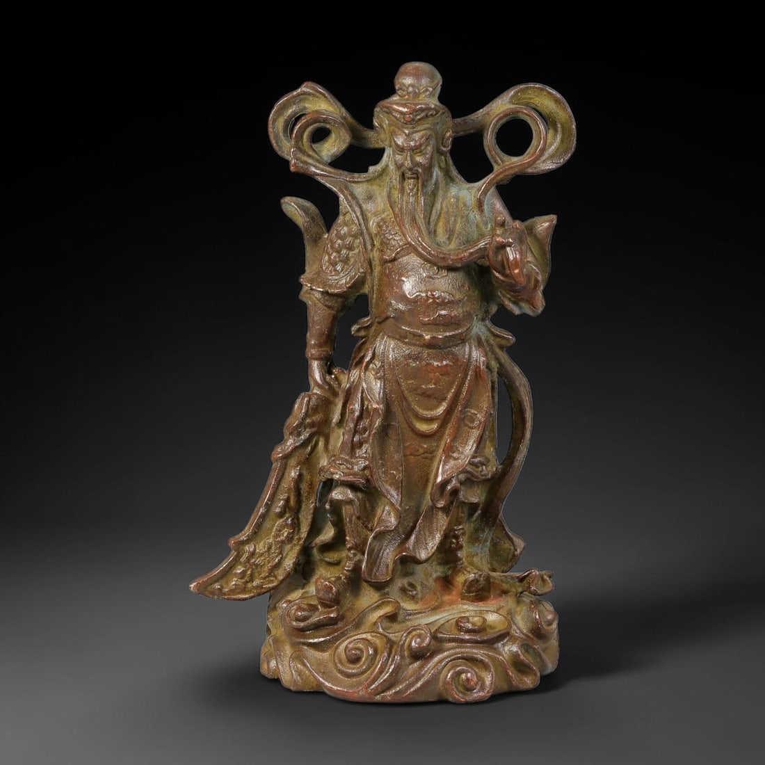 A Chinese Bronze Standing Figure of Guan Gong, 15AX: Cast bronze figurine depicting Guan Gong, standing atop swirling cloud base. The deity is clad in detailed armour and layered robes, with flowing ribbon elements framing the head, a long beard, and ho