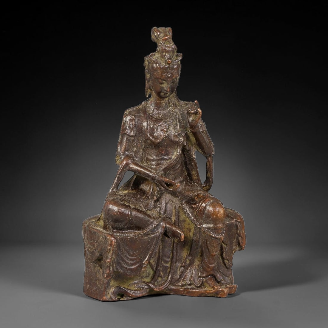 A Chinese Bronze Seated Guanyin Figure, 17AX: Cast bronze figure of Guanyin in the royal ease pose, seated upon a shaped rocky plinth. The deity wears layered flowing robes and an ornate headdress, with one hand raised in mudra and the other rest