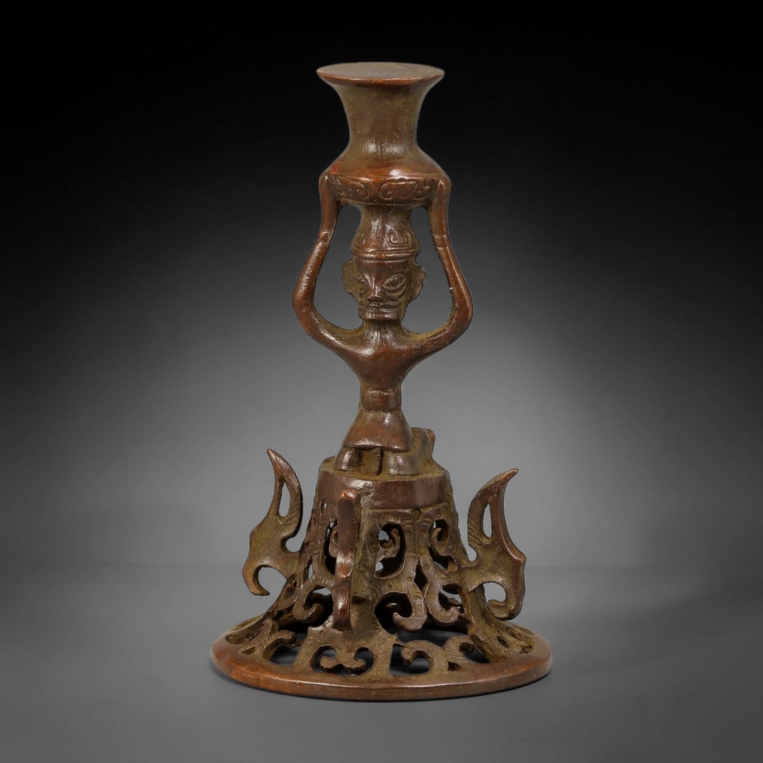 A Chinese Archaistic Bronze Figural Candlestick, 18AX: Cast bronze candlestick in archaistic style, featuring a central stylized humanoid figure supporting the upper candle holder. The circular base is decorated with elaborate openwork scroll and beast mo