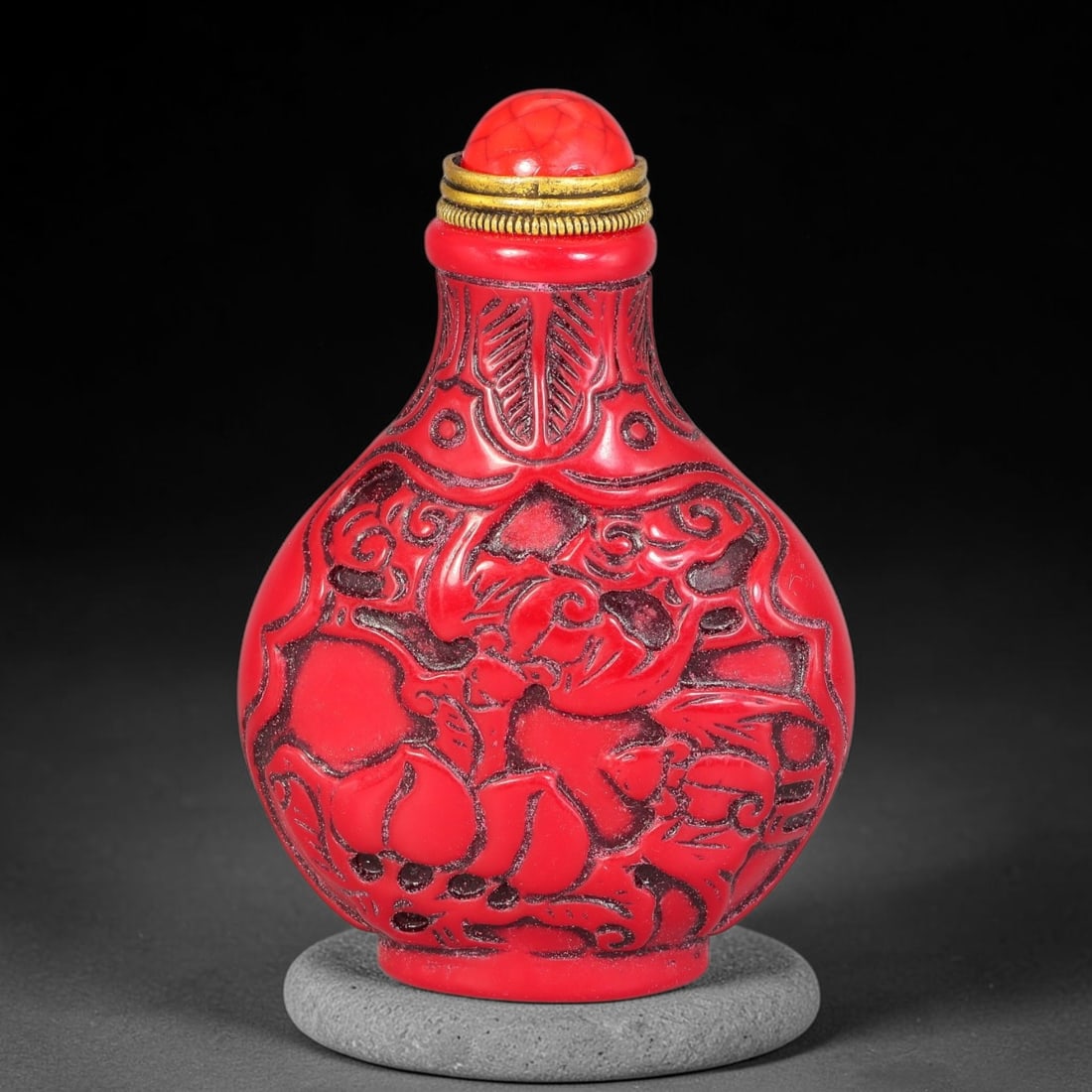 A Chinese Red Relief Carved Lotus and Figural Snuff Bottle, 92AV: Pear-shaped snuff bottle with vibrant red relief carving over dark recessed ground, depicting figures amid lotus blooms and cloud scroll ornamentation. The neck is decorated with stylized leaf pattern