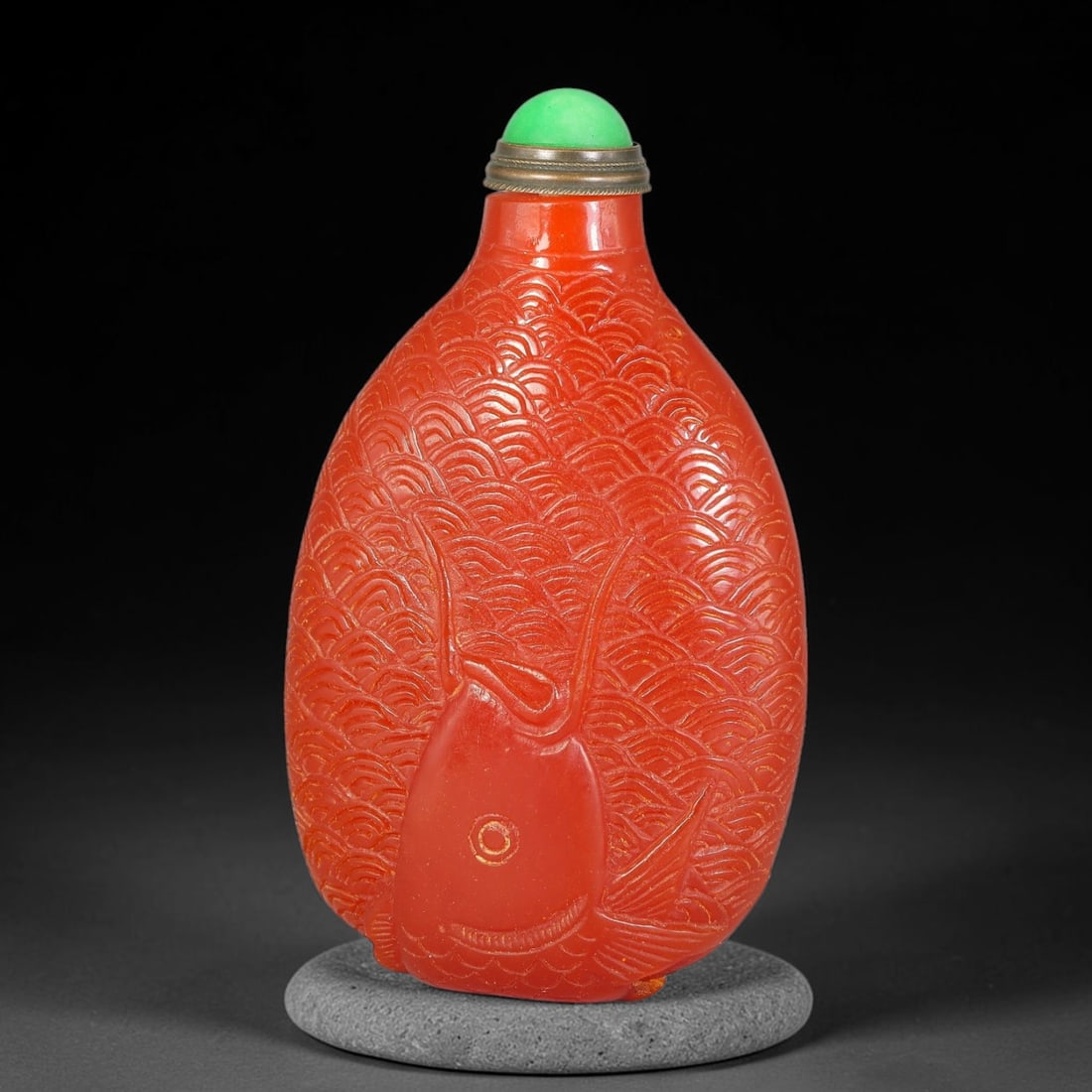 A Chinese Red Carved Fish Form Snuff Bottle, 83AV: Ovoid red snuff bottle finely carved in the form of a carp, with detailed overlapping scale and wave patterns covering the body. It is fitted with a green cabochon stopper set within a metal mount. He