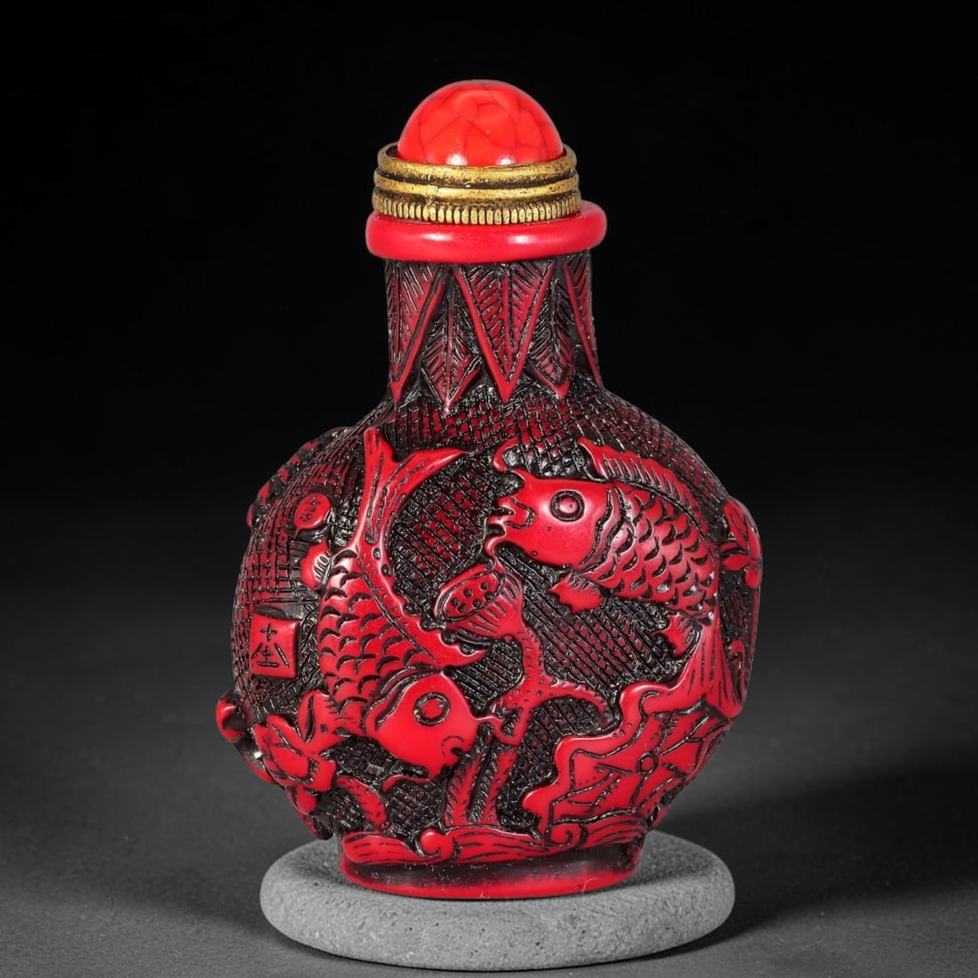 A Chinese Red Relief Carved Carp and Lotus Snuff Bottle, 3AX: Globular snuff bottle with bright red relief carving over dark recessed ground, decorated with paired carp swimming amid lotus leaves and seed pods, accented by seal characters. The neck bears pointed