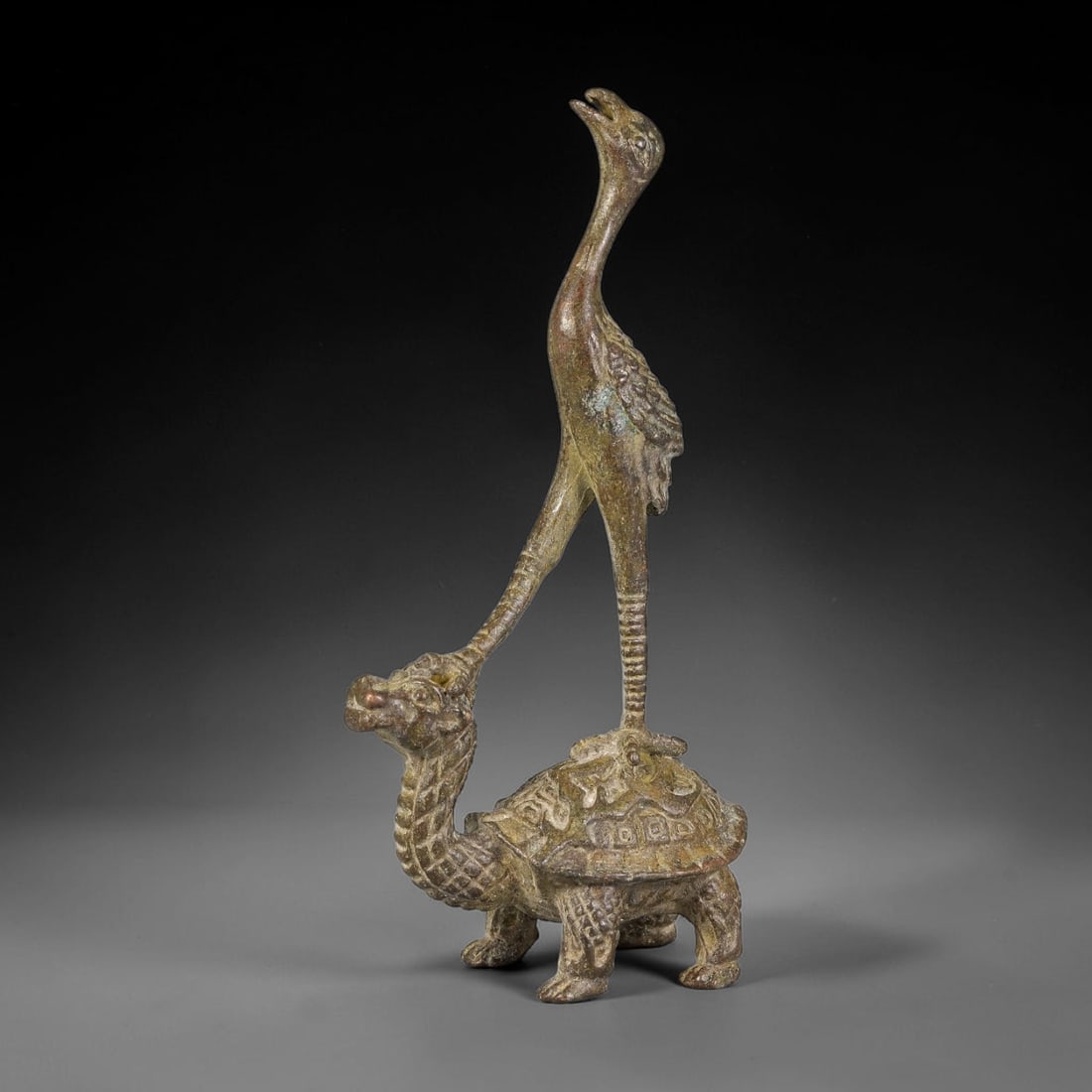 A Chinese Bronze Tortoise and Crane Figurine, 64AX: Cast bronze group depicting a crane standing atop a dragon tortoise, a traditional motif symbolizing longevity. The crane features an elongated neck, open beak and fine feather detailing; the dragon t