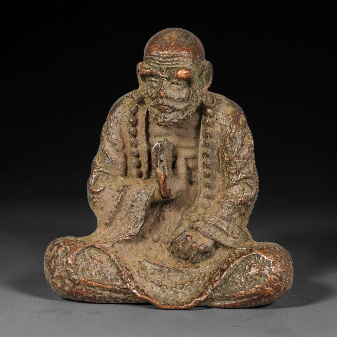 A Chinese Bronze Seated Luohan Figurine, 20AX: Cast bronze figure of a seated Luohan in meditative pose, with deeply wrinkled facial features, full beard, long prayer beads draped over the shoulders and textured robe detailing. The bronze displays