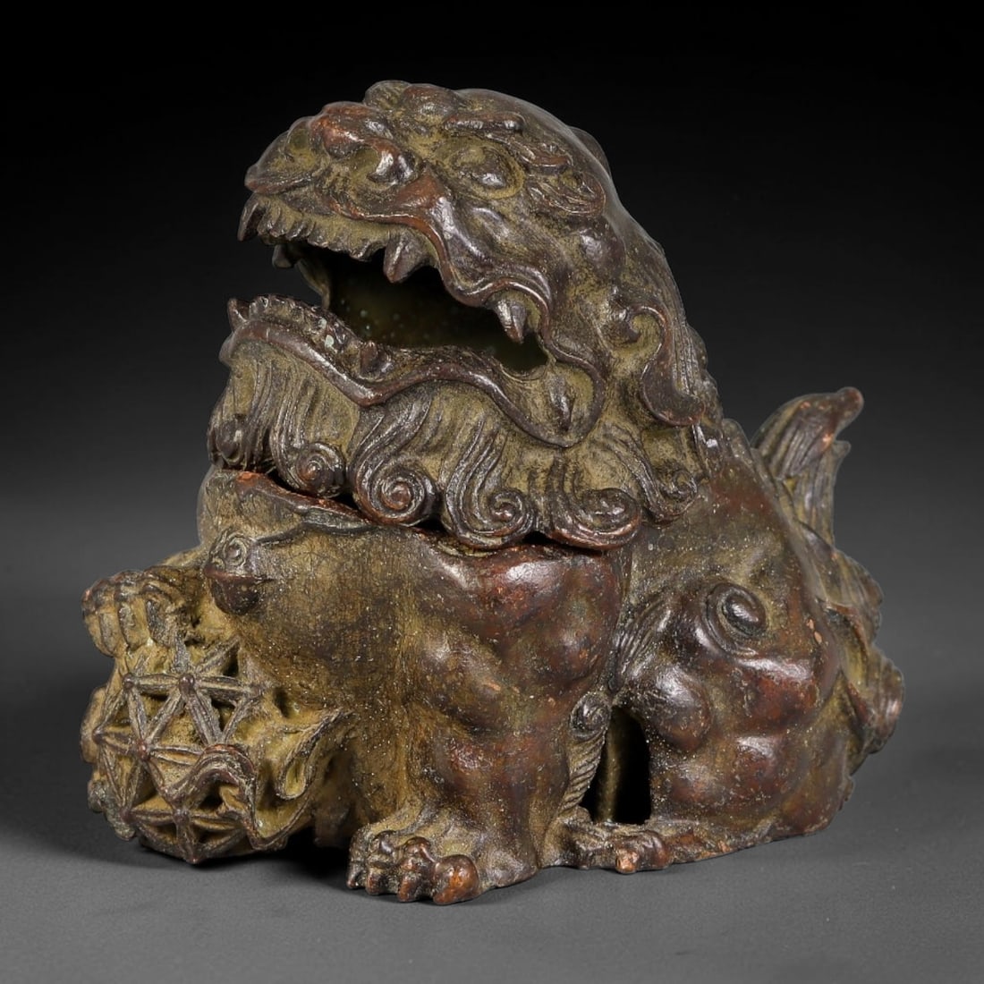A Chinese Bronze Fu Lion Split Form Censer with Pierced Ball, 56AX: Cast bronze censer modelled as a seated fu lion with hinged split head opening to form the incense chamber. The lion is detailed with curled mane, bared fangs and textured musculature, one paw resting