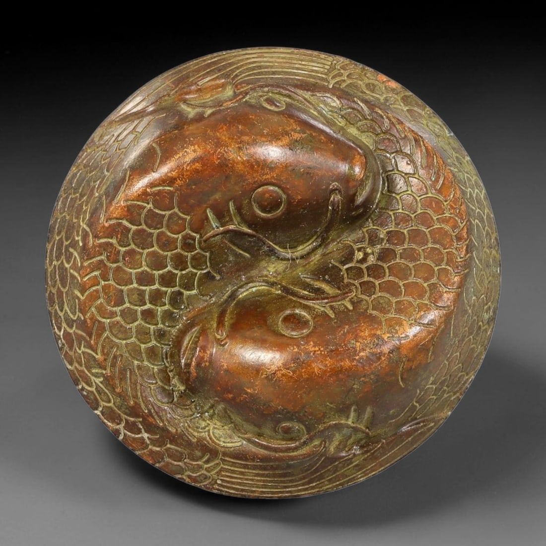 A Chinese Bronze Twin Fish Circular Paperweight, 36AX: Cast bronze circular paperweight with high relief twin fish motif encircled by engraved scale and wave detailing. The underside bears a square Qianlong reign mark. The bronze surface displays aged pat