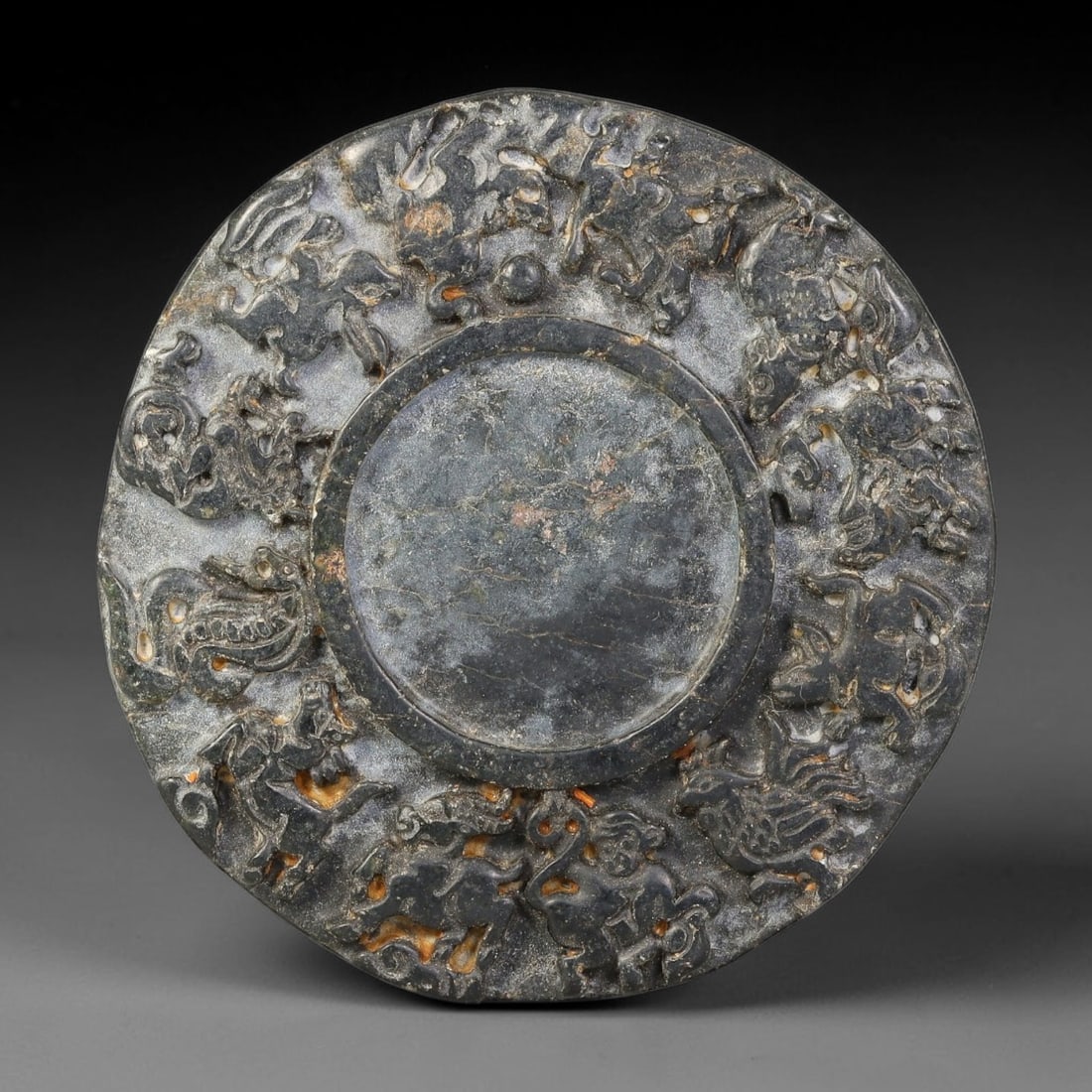 A Chinese Stone Zodiac Plaque, 47BD: Carved dark stone circular plaque with a raised flat central medallion. The surrounding field carries high-relief depictions of the twelve zodiac animal figures arranged in a continuous circular regis