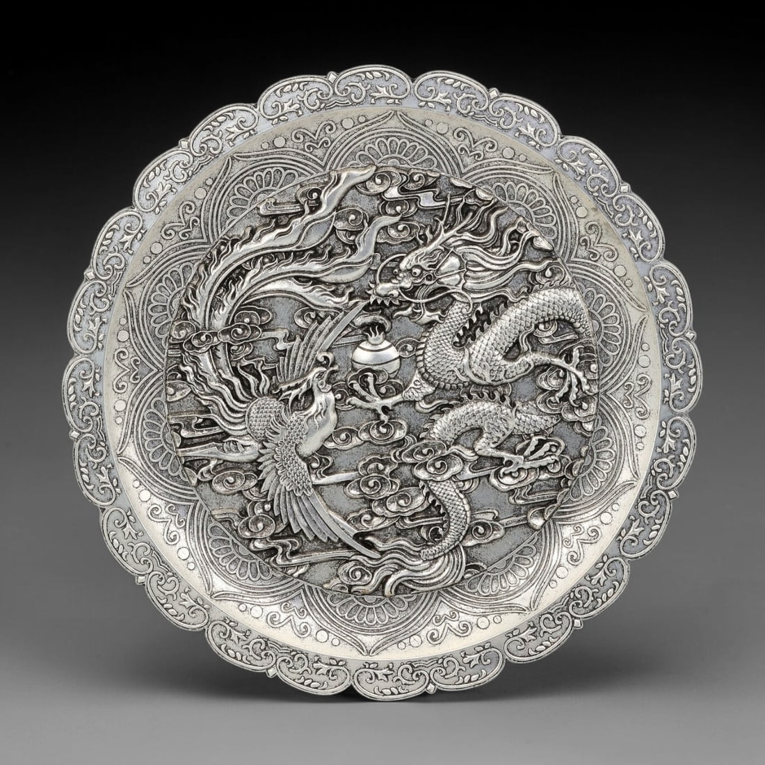 A Chinese German Silver Dragon and Phoenix Scalloped Dish, 44BD: Cast German silver dish with a scalloped flower-form rim. The central field carries high-relief dragon and phoenix figures confronting a flaming pearl among cloud scrolls. Concentric bands of stylized