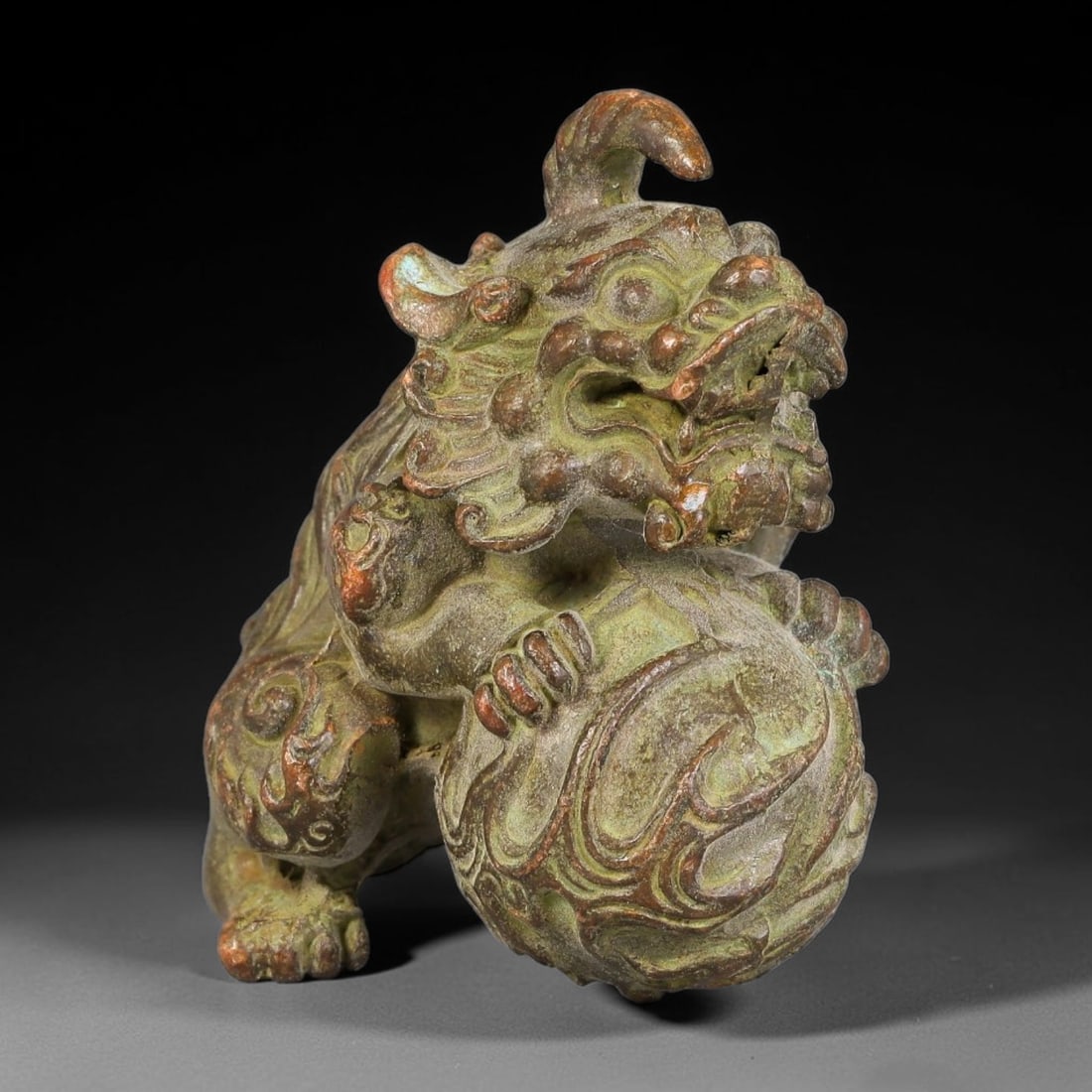 A Chinese Bronze Qilin Guardian Lion Playing with a Ball Effigy, 44AX: Cast bronze guardian lion depicted in a crouched pose with one paw resting on an ornately patterned ball, featuring layered curled mane details and expressive facial carving. The bronze surface has we
