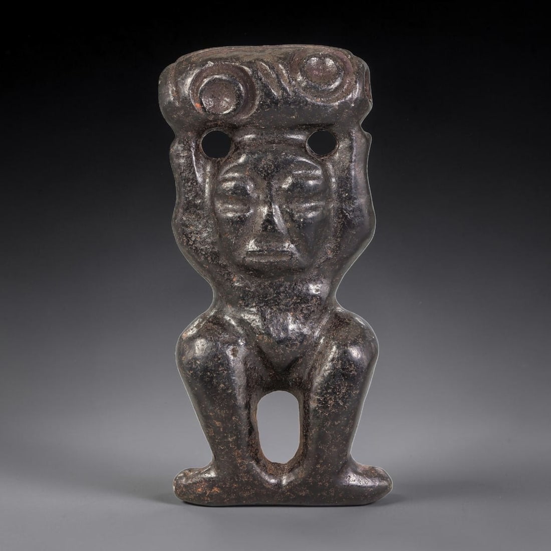 A Chinese Hematite Carved Anthropomorphic Figure, 34AX: Carved hematite anthropomorphic figure depicted in a squatting pose, hands raised to support a top element with circular motifs. Pierced openings are present on the shoulders and between the legs. The