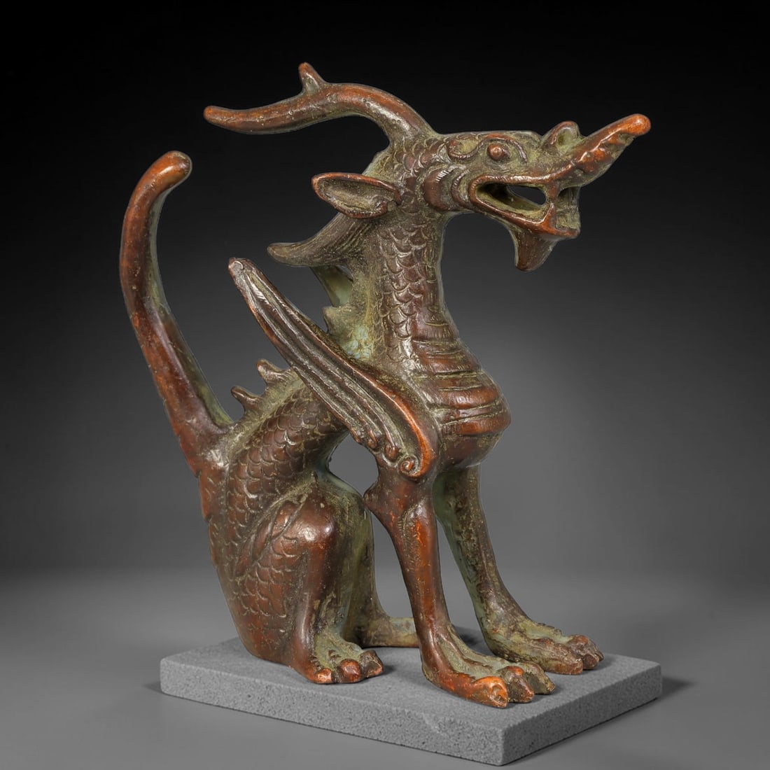 A Chinese Bronze Winged Dragon Figurine, 78AX: Cast bronze seated winged dragon with antlered horns, open mouth, carved scale patterning covering the torso, stylized wing details and curved raised tail. The bronze has a deep reddish-brown aged pat